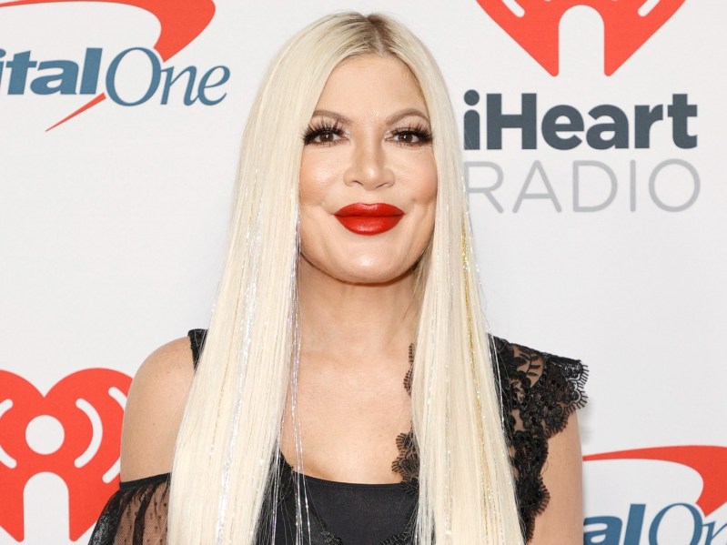 Tori Spelling smiles while wearing a black dress. She has her platinum blonde hair striaghtened and parted down the middle, and is wearing bright red lipstick