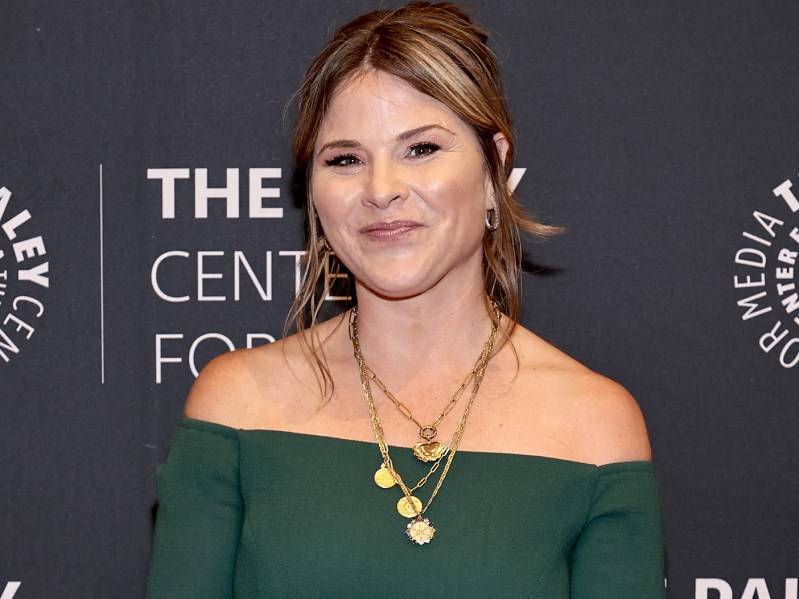 Jenna Bush Hager smiles against a charcoal gray background. She is wearing a deep forest green, off-the-shoulder dress with a gold necklace. Her hair is pulled back into a low bun.