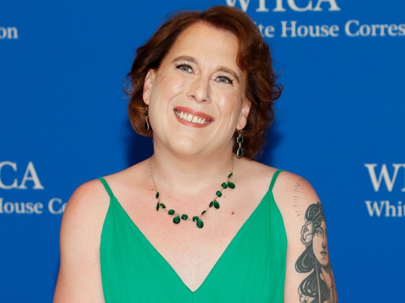 Amy Schneider grins against a royal blue background. She is wearing a green dress with spaghetti straps and a beaded necklace. Her right arm has a large tattoo on it.