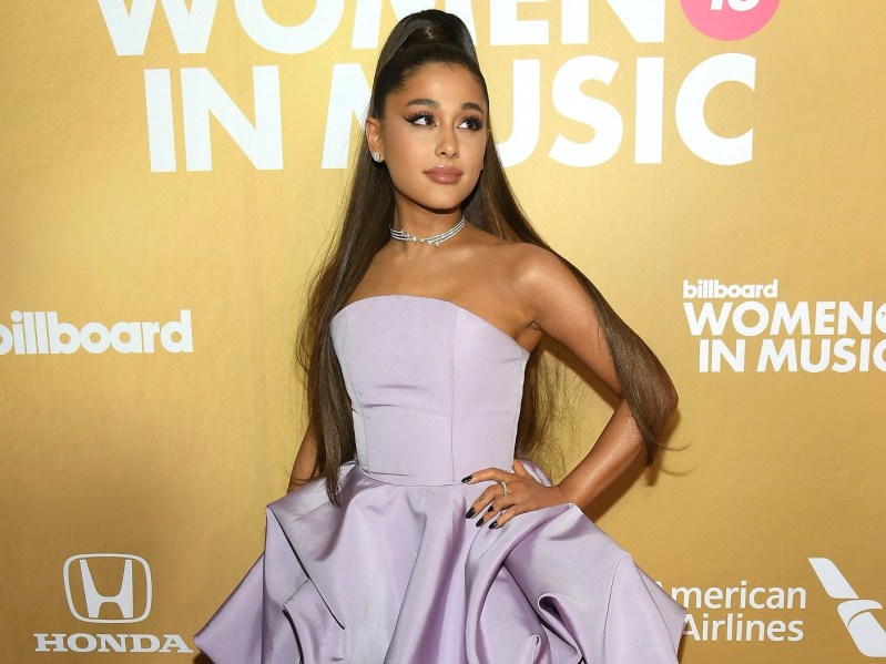 Ariana Grande standing against yellow background. Her hand is on her hip and she is looking off to the viewer's right. She is wearing a strapless, lavender gown and her hair is half-up