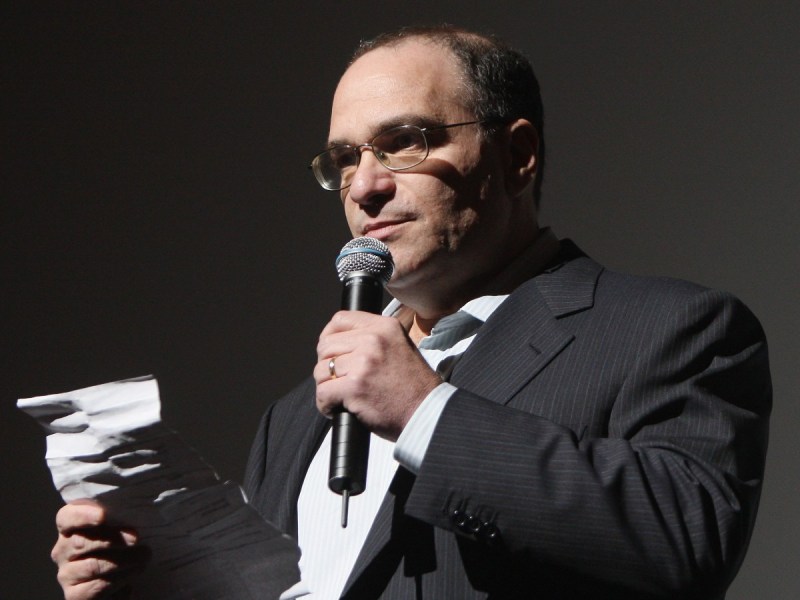 Bob Weinstein standing with microphone and holding piece of paper. He is wearing a white button-down shirt with a black blazer
