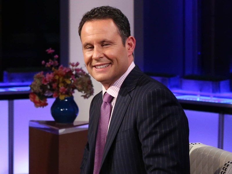 Brian Kilmeade smiles at the camera. he is wearing a white shirt, black suit jacket, and a purple tie. There is a vase of flowers sitting on a platform behind him