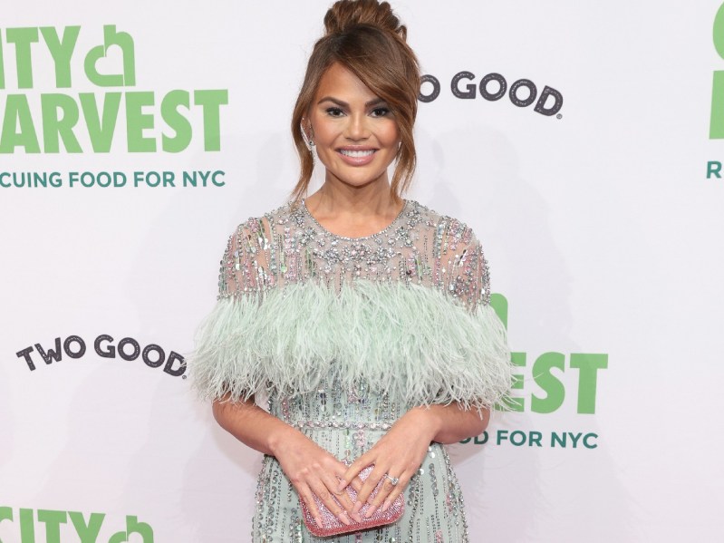 Chrissy Teigen wears a green and silver dress on the red carpet against a white background