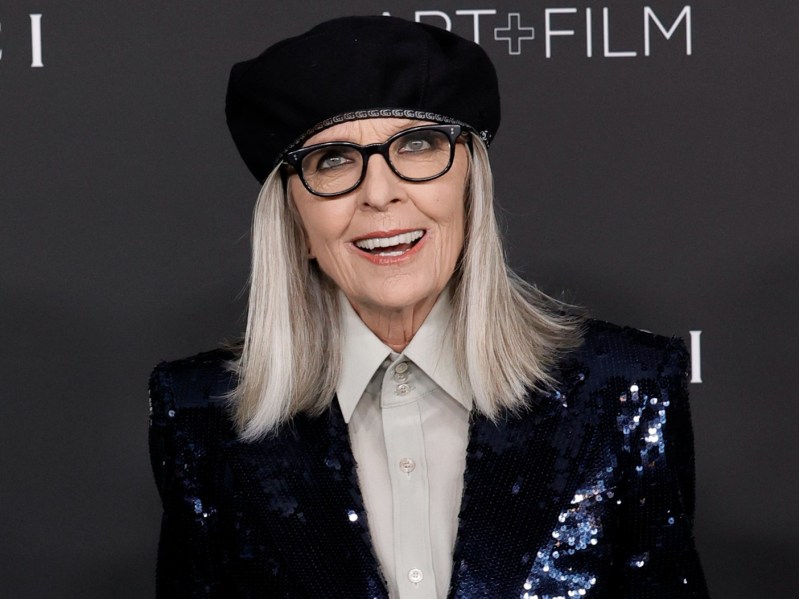 Diane Keaton smiles while wearing a black sequined suit jacket over a gray shirt and black hat