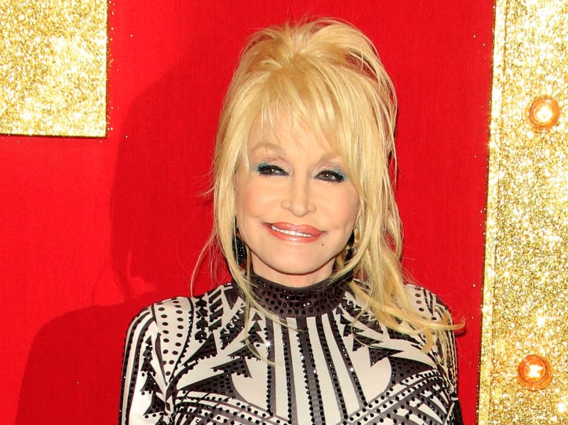 Dolly Parton wears a gown with a black pattern against a red and gold background on the red carpet