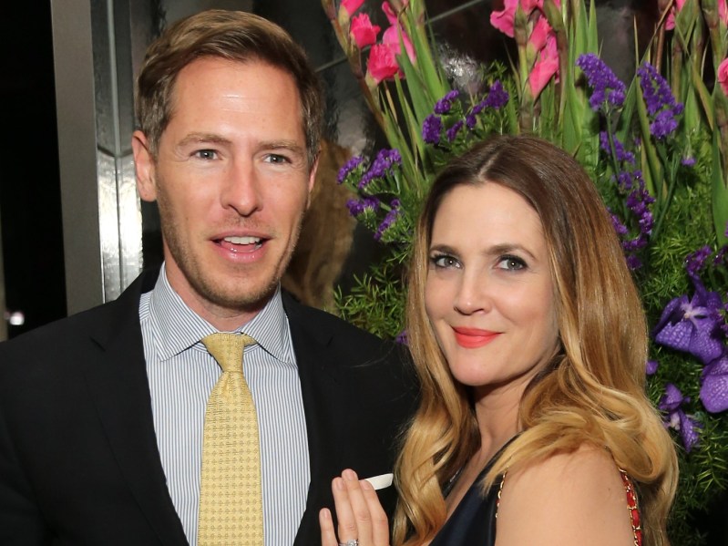 Will Kopelman (L) wearing suit with yellow tie standing next to Drew Barrymore (R) wearing black dress and red lipstick