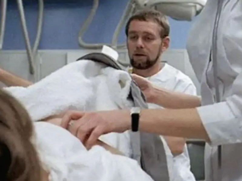 A man with blonde hair and a beard wearing medical scrubs is seen tending to a patient. We cannot see the patient in the photo