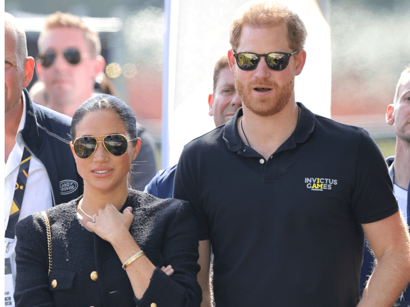 Meghan Markle (L) wearing black top and sunglasses with hair pulled back, walking outside next to Prince Harry (R) wearing navy Invictus Games polo and sunglasses