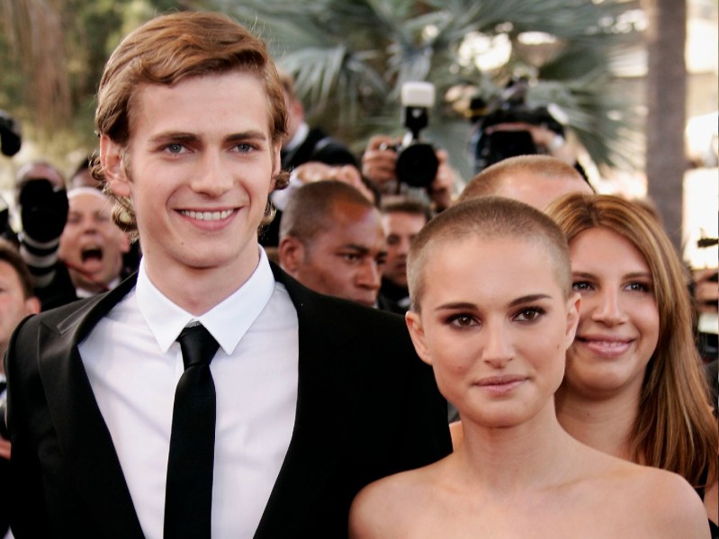 Hayden Christensen, wearing a black suit, stands with Natalie Portman, sporting a shaved head and a strapless black gown, on the red carpet