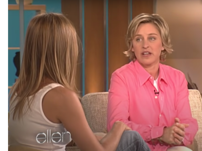 Ellen Degeneres (R) is wearing a bright pink button-down top and looking to an interviewee, whose face we cannot see but who is wearing a white tank top and has long, blonde hair