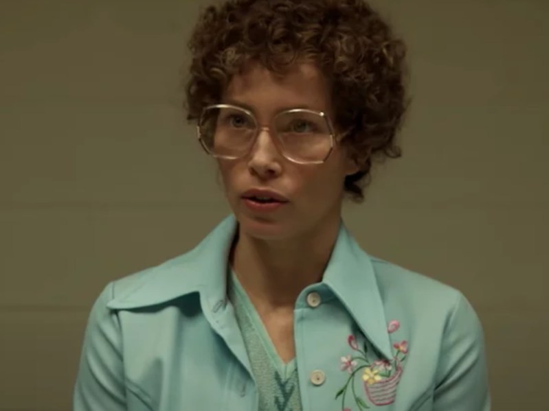 Jessica Biel is dressed as Candy Montgomery. She is wearing a curly, short wig and large glasses with a light blue shirt/jacket combo