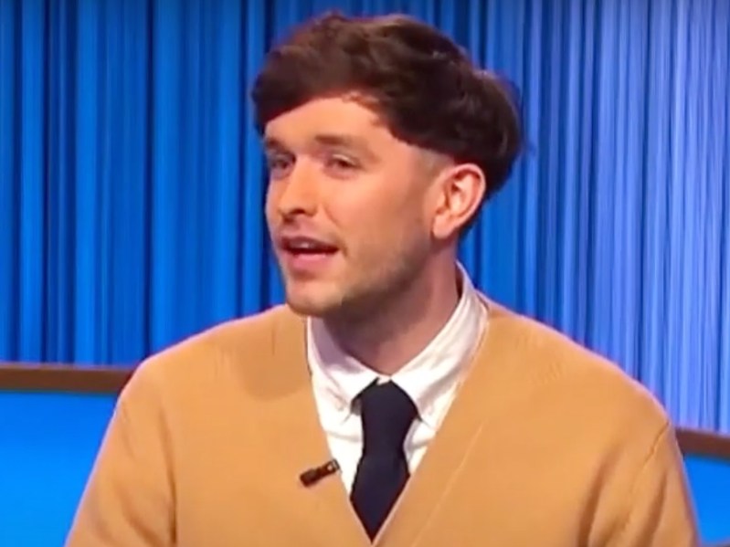 Julian Glander is wearing a white shirt and black tie with a ten/gold cardigan. He appears to be speaking