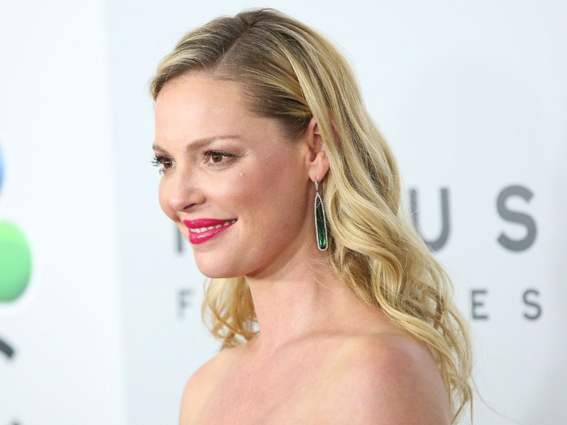 Katherine Heigl from the shoulders up, smiling in profile with red lips and hair down and curled.