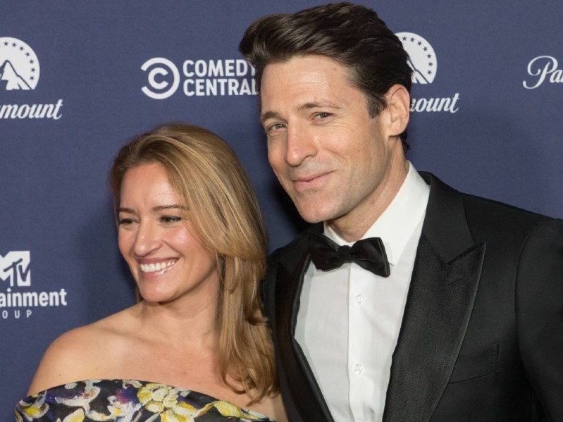 Katy Tur (L) wearing off-shoulder black and yellow patterned dress, standing next to her husband, who is wearing a white button down shirt with a black suit jacket and black bow tie