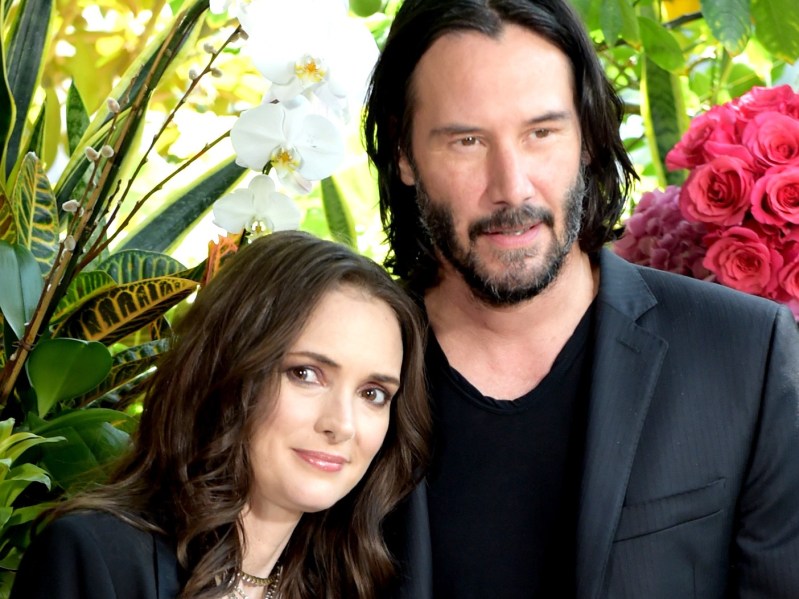 Winona Ryder wears a dark blazer over a dark shirt and leans against Keanu Reeves, also in a dark blazer and black shirt, in front of indoor plants