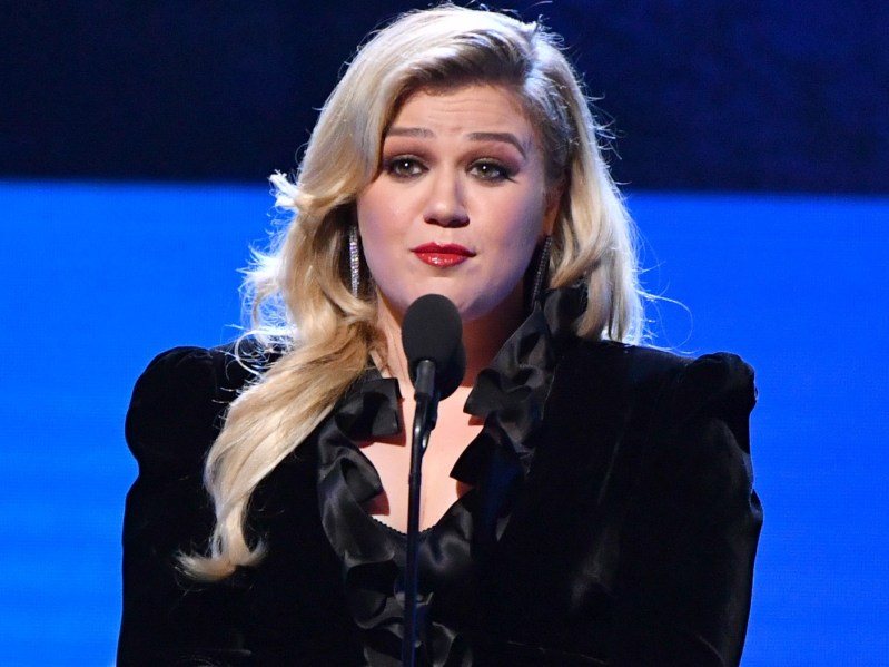 Kelly Clarkson speaking into a microphone. She is wearing an all-black suit and her blonde hair is down and curly.