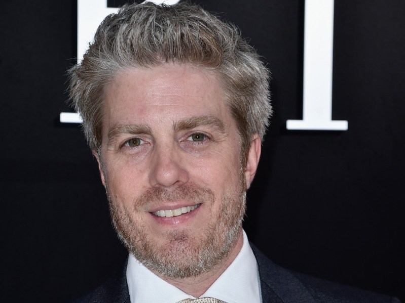 Closeup of Kyle Eastwood smiling with white collared shirt and black jacket against a black background with white letters on it