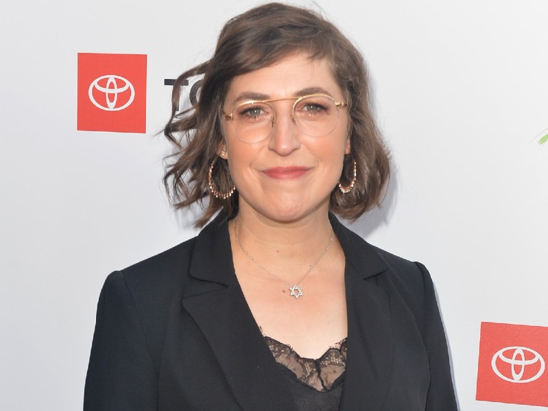 Mayim Bialik smiles while wearing gold-rimmed glasses and a black blazer over a black, lace shirt