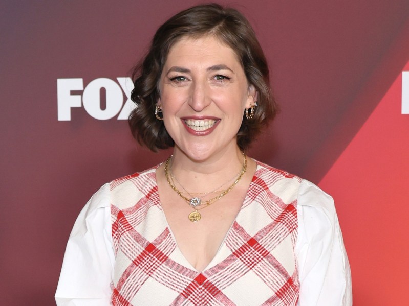 Mayim Bialik smiles while wearing a white and red plaid dress. Her hair is in a bob haircut