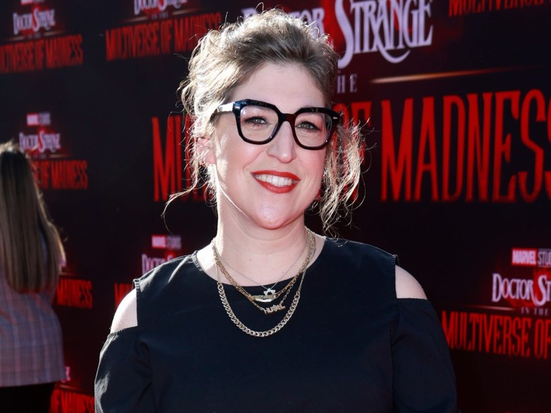 Mayim Bialik wearing black, cold-shoulder dress with gold necklace. her hair is pulled back into a bouffant and she is wearing thick-rimmed black glasses