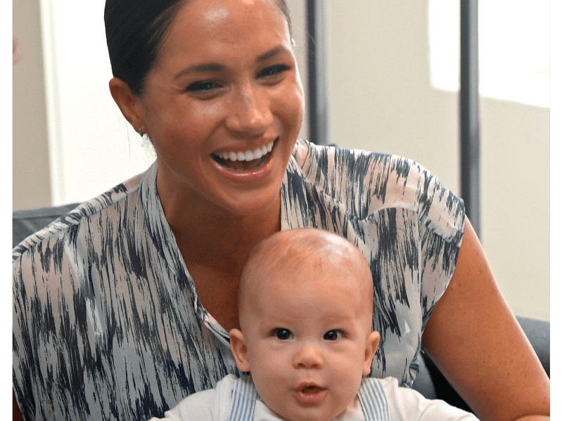 Meghan Markle smiling with her hair pulled back wearing a patterned dress. In her lap is her infant son Archie, with a quizzical look on his face