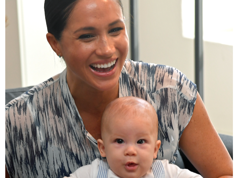 Meghan Markle smiling with her hair pulled back wearing a patterned dress. In her lap is her infant son Archie, with a quizzical look on his face