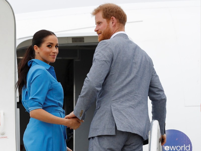 Meghan Markle, in a blue dress, walks with Prince Harry, in a gray suit, up airplane stairs during their tour of Tonga