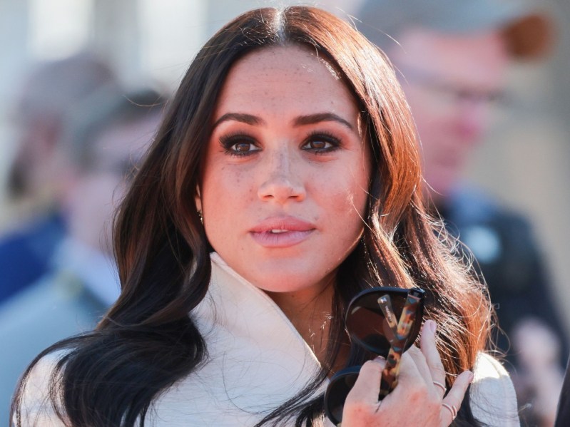 Closeup of Meghan Markle looking into the distance. She is holding a pair of sunglasses in her hand.