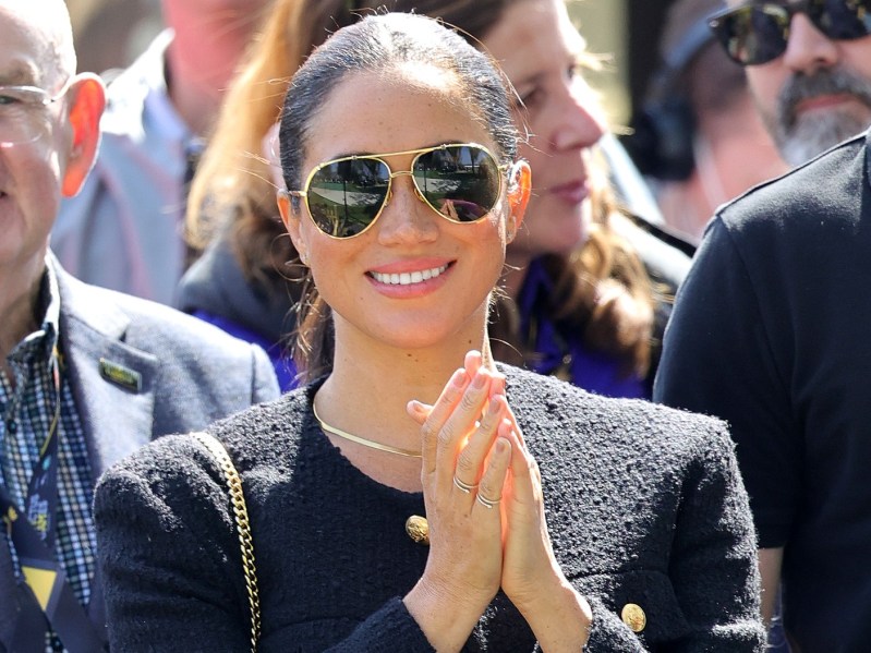 Meghan Markle smiles with her hands clasped. She is outside and has on a navy blue jacket with gold buttons and trim. She has her hair pulled back and is wearing aviator-style sunglasses.