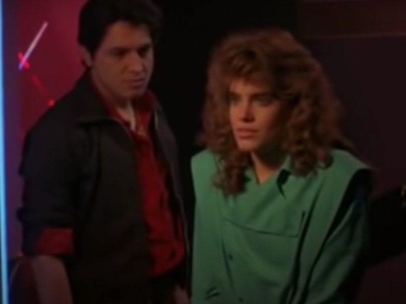 A dim still from a movie shows a man with black hair and a leather jacket looming behind a girl with large curls and a green top