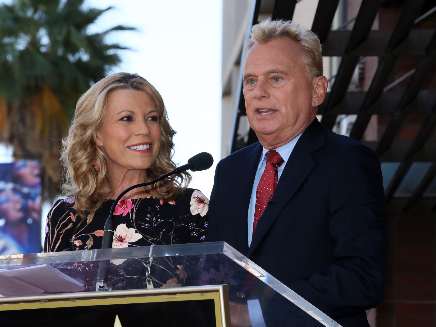 Vanna White (L) wearing a floral dress tanding next to Pat Sajak (R) wearing black suit with red tie, speaking into microphone.