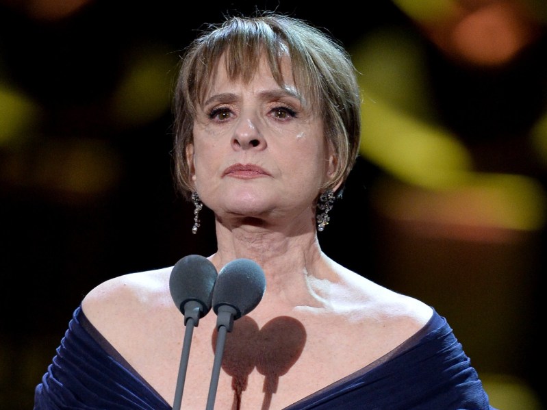 Patti Lupone with a straight face standing in front of two microphones. She is wearing an off-the-shoulder, deep blue gown.