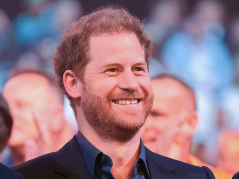 Prince Harry sits in a crowd and smiles, wearing navy button-down