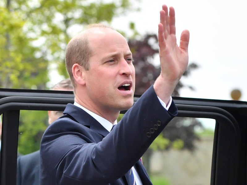 Prince William holds his hand up and appears to be flagging someone down. He is wearing a deep navy blue suit jacket over a white dress shirt.