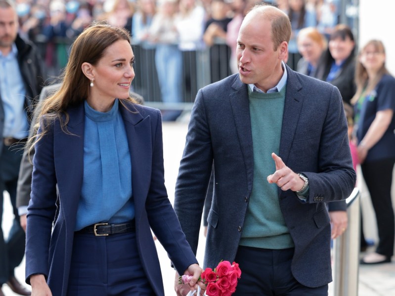 Kate Middleton (L) wearing blue top and darker blue jacket, walking next to Prince William (R), wearing green sweater and dark navy jacket. Middleton is holding shopping bags