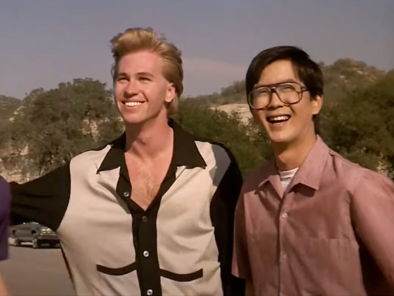 Two young men stand side-by-side outdoors. One has blonde hair and is wearing a black and cream colored button-down. The other has dark hair and blacj glasses, and is wearing a salmon/mauve-colored button-down. Both men are smiling off into the distance.