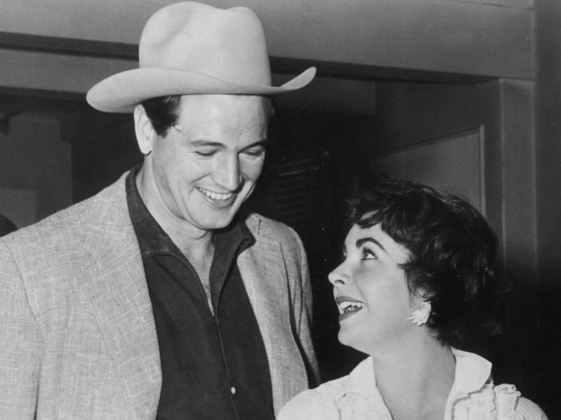 (Black and white photo): Rock Hudson, dressed in a shirt and blazer combo with a large cowboy hat, smiles at Elizabeth Taylor, who is looking back at him while cutting cake.