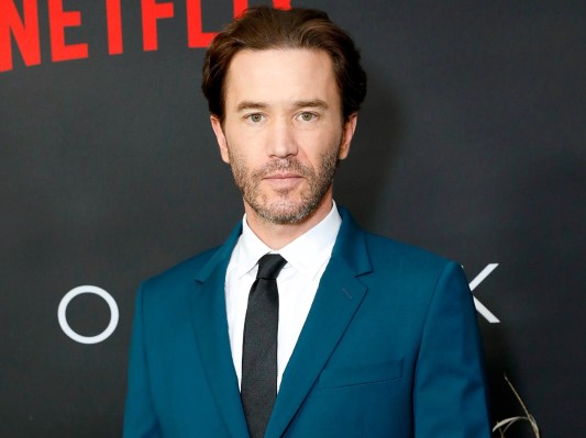 Tom Pelphrey looks directly at the camera. He is wearing a white shirt, black tie, and deep blue suit jacket.