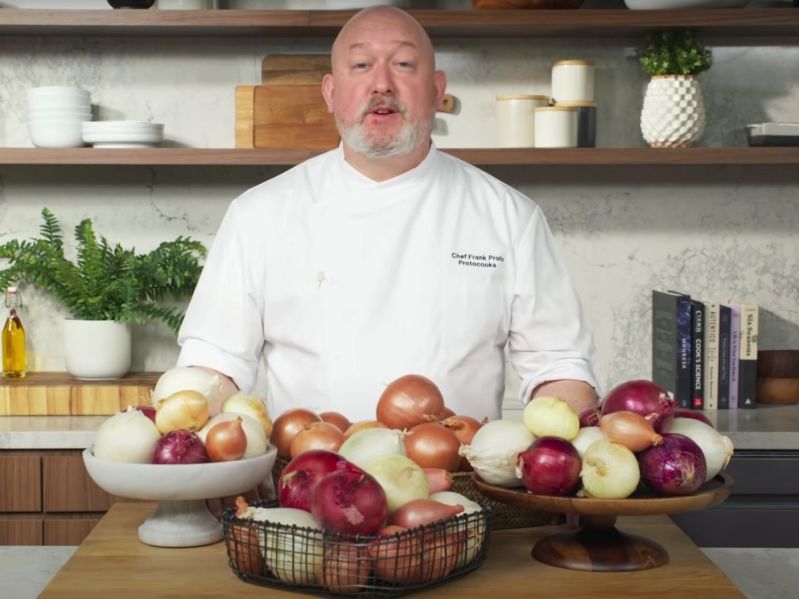 Chef Frank behind a table filled with onions in his video on best methods to peel and cut onions