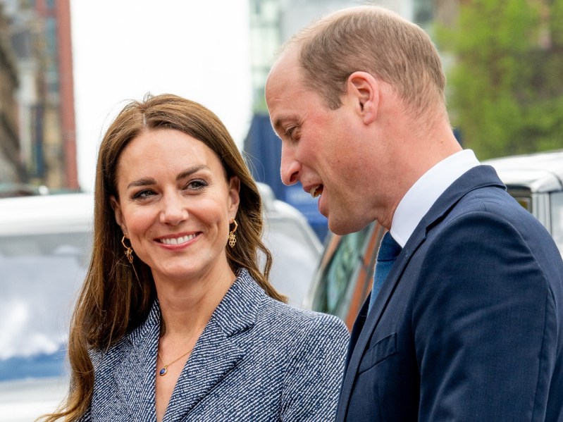 Kate Middleton (L) is wearing a grey-blue suit jacket and smiling at Prince William (R), who is standing in profile and appears to be speaking. William is wearing a navy blue suit jacket with a white collared shirt.