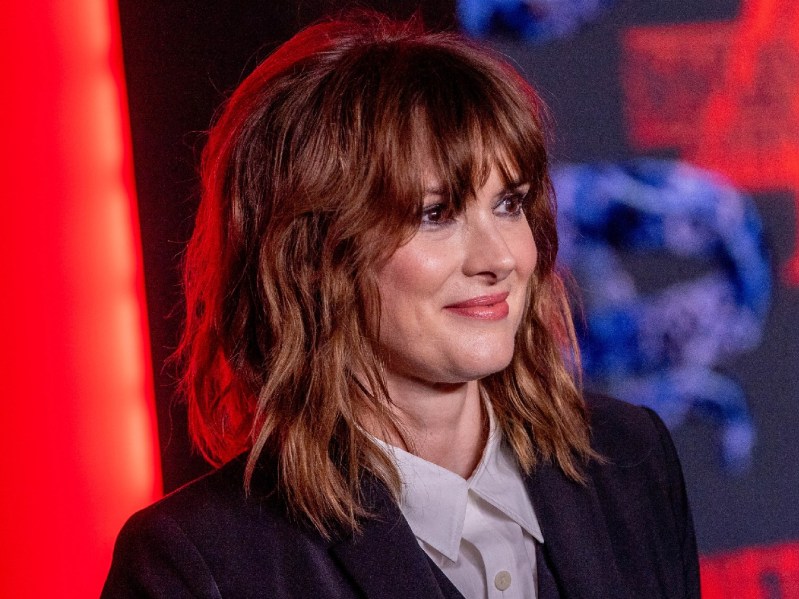 Winona Ryder wearing grey shirt and black blazer. She has blunt bangs and her brown hair is shoulder-length