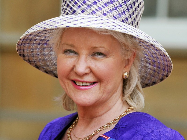 Angela Kelly smiles while wearing a purple dress and purple and white plaid hat