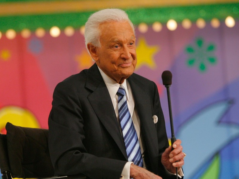 Bob Barker smiles while wearing a black suit jacket over a white dress shirt and blue striped tie. He is holding a microphone