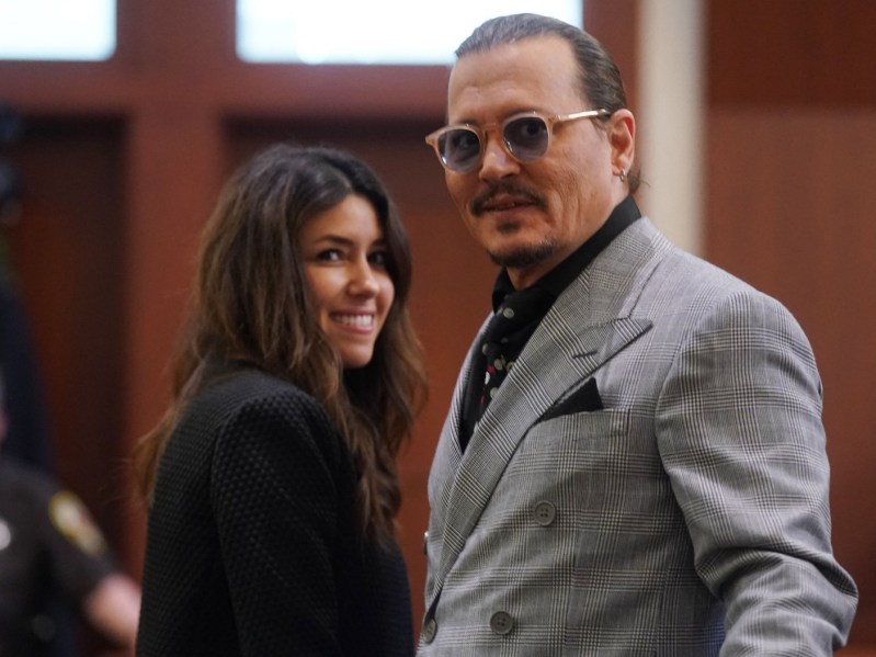 Camille Vasquez (L) smiling in a black blazer, standing next to Johnny Depp, who is also smiling and wearing a gray suit jacket over a black shirt and patterned tie