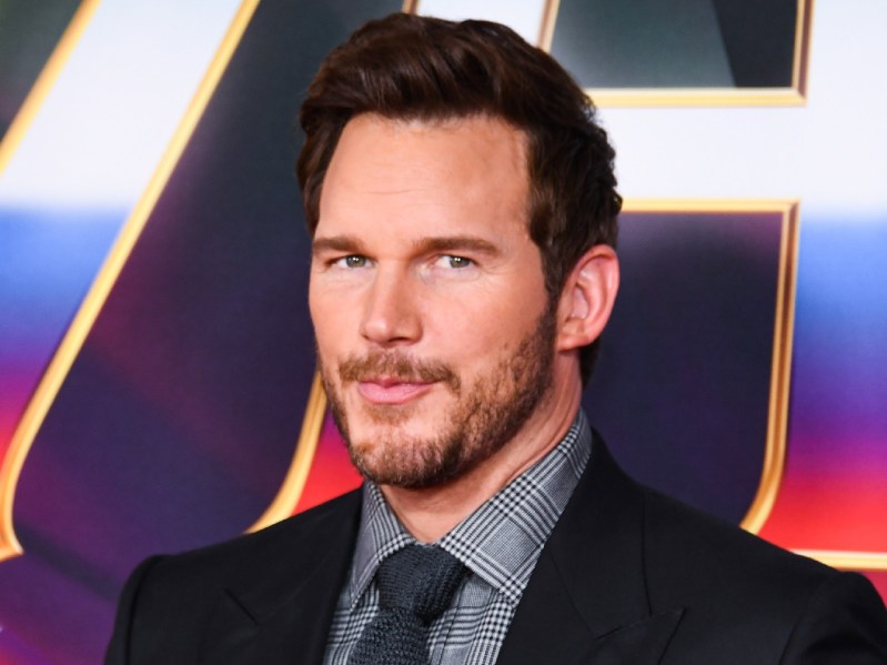 Chris Pratt looks at the camera while wearing black suit and tie over dark gray dress shirt