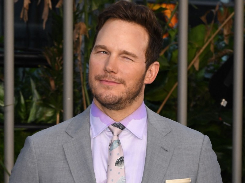 Chris Pratt winks while wearing a purple button down/tie and gray suit jacket