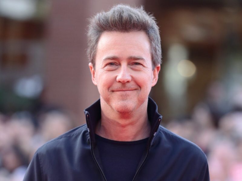 Edward Norton smiles while wearing ablack top and black zip-up jacket