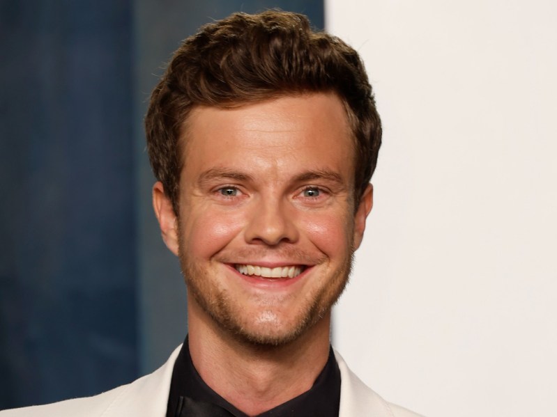 Closeup of Jack Quaid smiling wearing black and white suit
