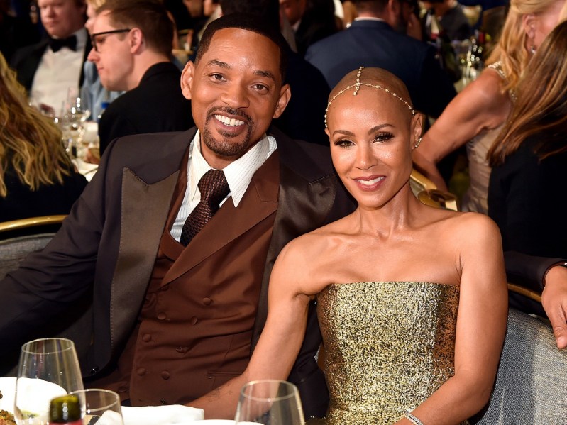 Will Smith, in a brown suit, sits with wife Jada Pinkett Smith, in a gold dress and headpiece, at a table indoors