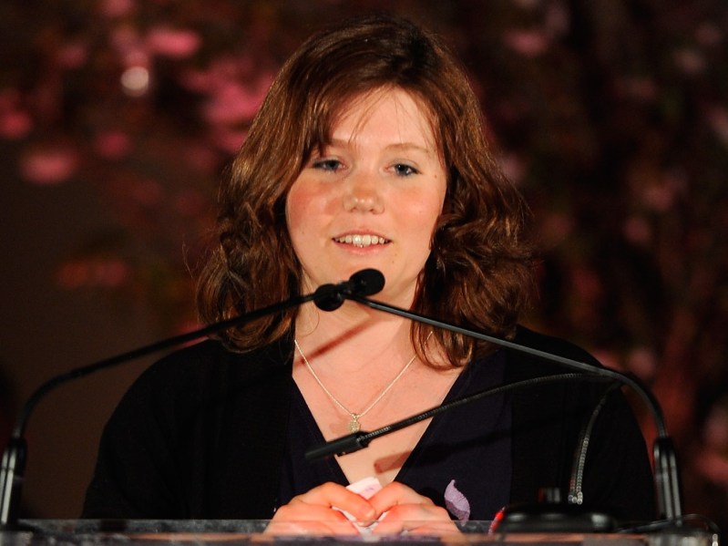 Jaycee Dugard stands at a podium and speaks into two microphones. She is wearing a black shirt and stands against a dark red, patterned backdrop